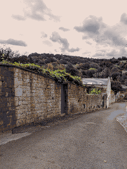 Looking down a road on a hill, following a bricked wall with plants growing over the top and inset garden gates. Trees from a nearby woodland peak above the wall as they rise back up the hill on the other side.