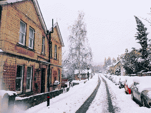 Car tyre tracks melt the snow that has blanket an urban street, a yellow stone house sits on the left and parked cars line the right.
