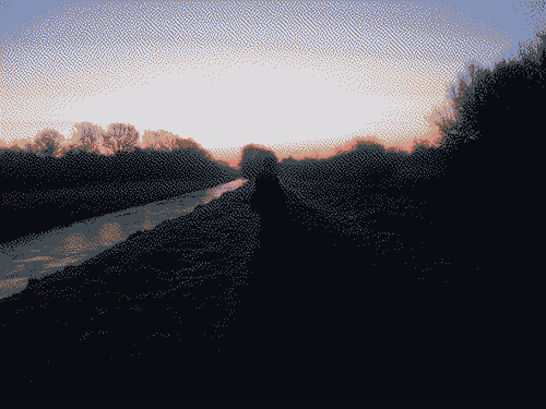 A orangey pink sun set with a river and trees in the foreground, dimly lit.