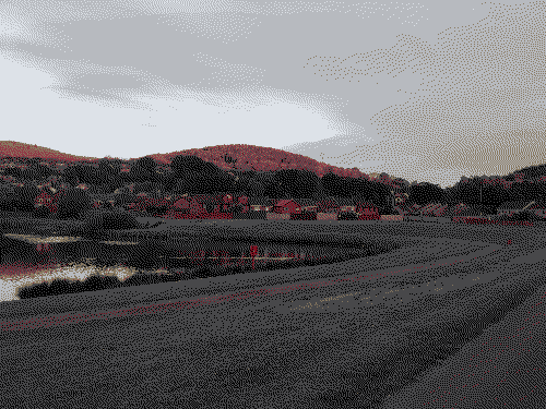A grass field wraps around a lake, with trees lining the edge, undulating hills in the background are lit up orange during the golden hour.